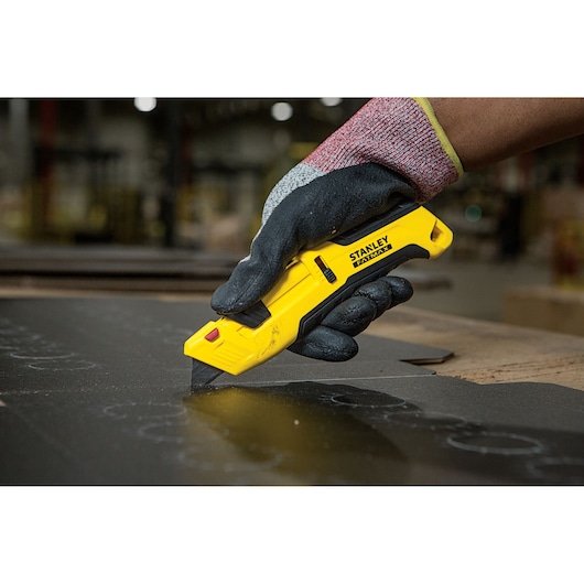 Photo of STANLEY FMHT10365-0 utility knife in a gloved hand cutting black material, indoor workshop setting.