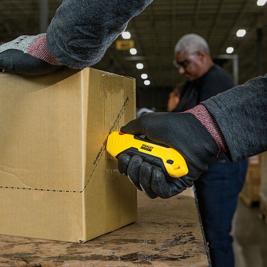 Stanley FMHT10369 utility knife cutting cardboard box, close-up, worker wearing black gloves.