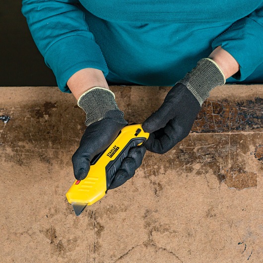 STANLEY FMHT10369 utility knife held in gloved hands on a workbench, yellow and black body, top view.