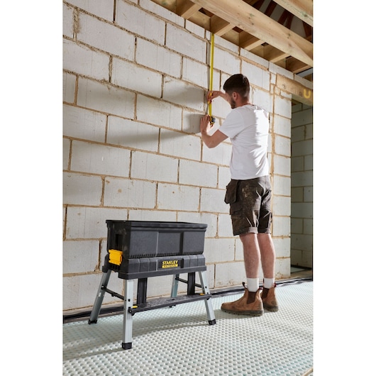 STANLEY FMST81083-1 black foldable sawhorse tool box on site, construction worker measuring wall.