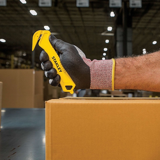 STANLEY STHT10355 yellow safety box cutter held by gloved hand cutting cardboard, warehouse background.