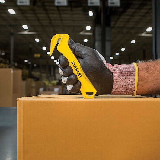 Stanley STHT10355 yellow safety cutter held by a gloved hand slicing a cardboard box in a warehouse.