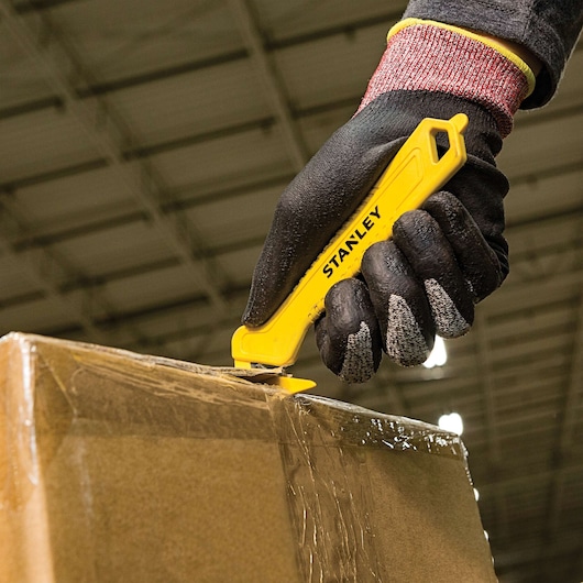STANLEY STHT10355 yellow safety utility knife cutting tape on a cardboard box, held by a gloved hand.