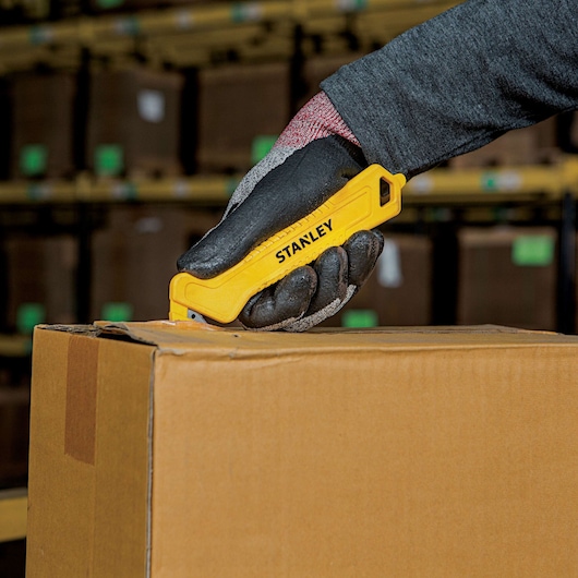 Photo of STANLEY STHT10355 yellow safety utility knife cutting cardboard box in warehouse setting.