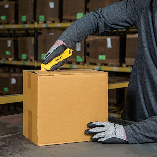 STANLEY STHT10355 utility knife cutting a cardboard box, worker wearing gloves, warehouse background.