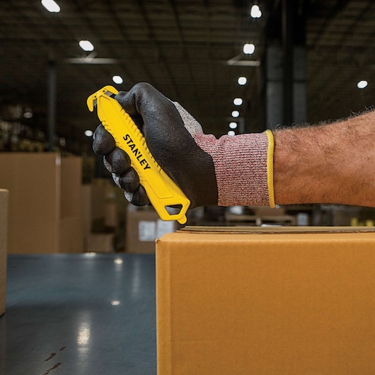 Stanley STHT10359 safety knife used to cut a cardboard box, shown in a warehouse setting.