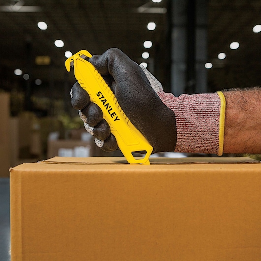 Stanley STHT10359 safety knife in gloved hand cutting cardboard box, close-up, warehouse background.