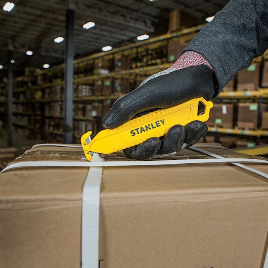 Stanley STHT10359 safety knife used to cut box straps, warehouse background, product held by gloved hand.