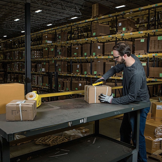STANLEY STHT10359 utility knife cutting box on warehouse table with shelves of packages in the background.