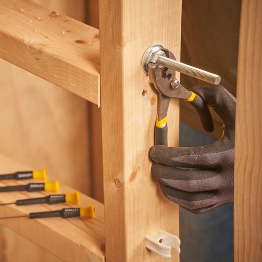 STANLEY STMT0-74101 pliers gripping a bolt on wooden frame, close-up photo, hand in black glove.