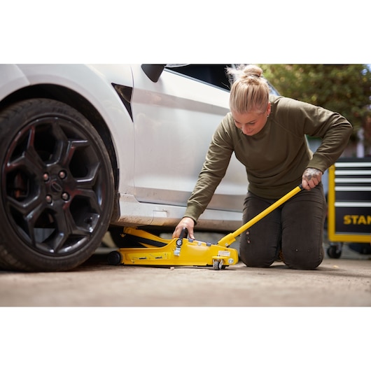 STANLEY STMT81251-1 yellow floor jack in use lifting a car, user kneeling beside, garage setting.